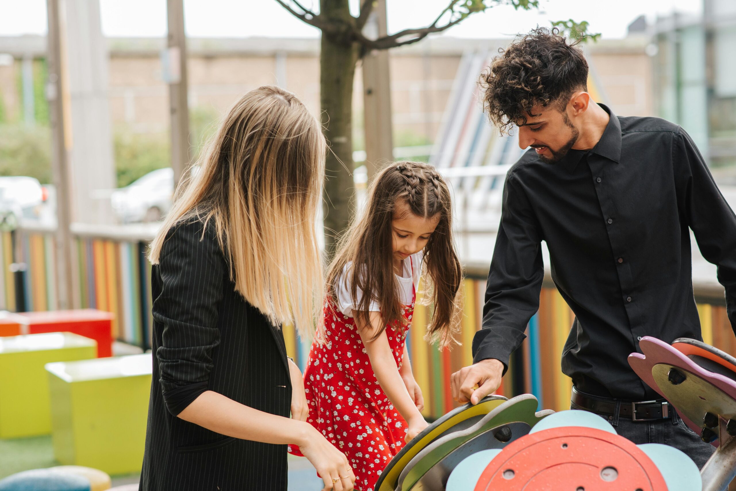 Little girl playing on playground with support of mother and father in casual clothes in summer day